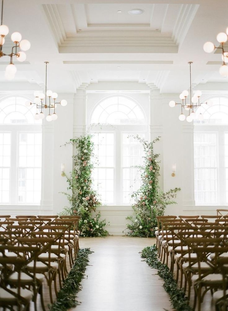 an aisle lined with rows of wooden chairs covered in greenery next to two large windows