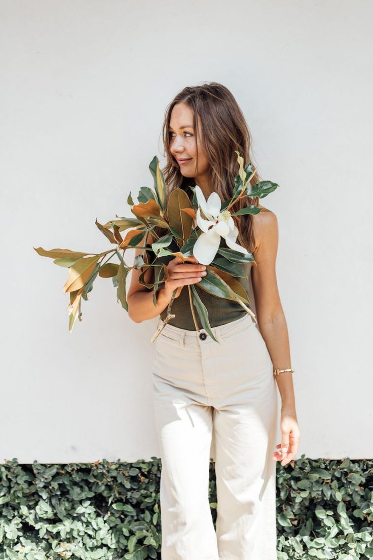 a woman holding flowers in her hands and smiling at the camera while standing against a wall