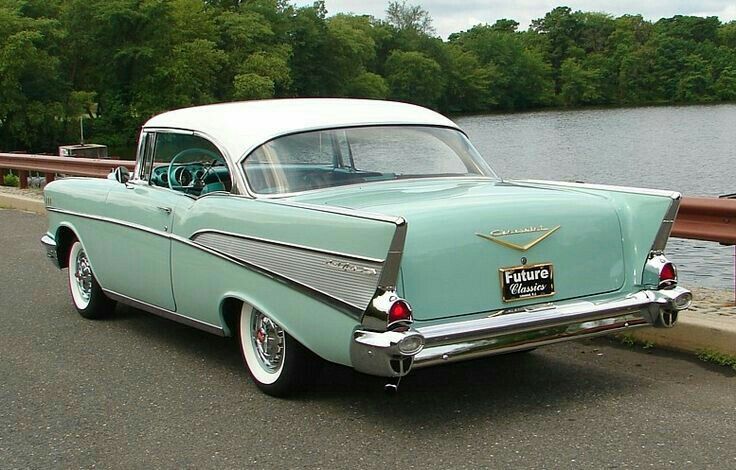 an old car is parked on the side of the road next to the water with trees in the background