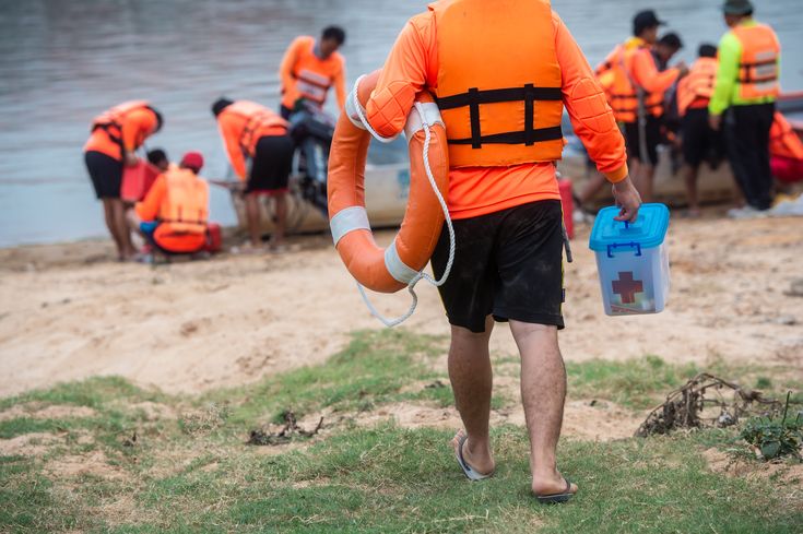 a man in an orange life jacket is carrying a blue bucket and some other people are standing on the shore