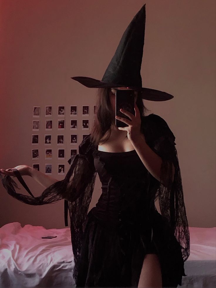 a woman in a witch costume taking a selfie with her cell phone while sitting on a bed