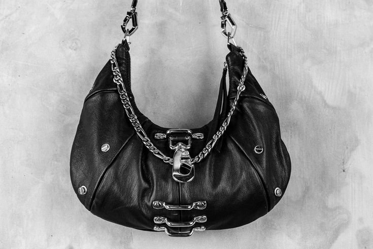 An ode to the timeless partnership of leather and chains, this leveled up black hobo bag is made from soft, premium genuine black leather and features a top shelf arrangement of silver hardware throughout.A centered, weighty metal industrial clip takes center stage, accompanied by an array of high contrast chromed out nailed rivets, centered feed through bars, and a draped top chain that doubles as a shoulder strap.Complete with a removable/adjustable leather and chain strap that can easily convert this purse from a shoulder bag to a comfortable crossbody bag, this design is an ode to the tough and timeless partnership of leather and chains.Bag is approx 13.5" x 7"Removable strap can extend up to 40" Black Bag Silver Hardware, Black Hobo Bag, Shelf Arrangement, Holster Bag, Convertible Crossbody Bag, Draped Top, Black Leather Purse, Top Shelf, High Contrast