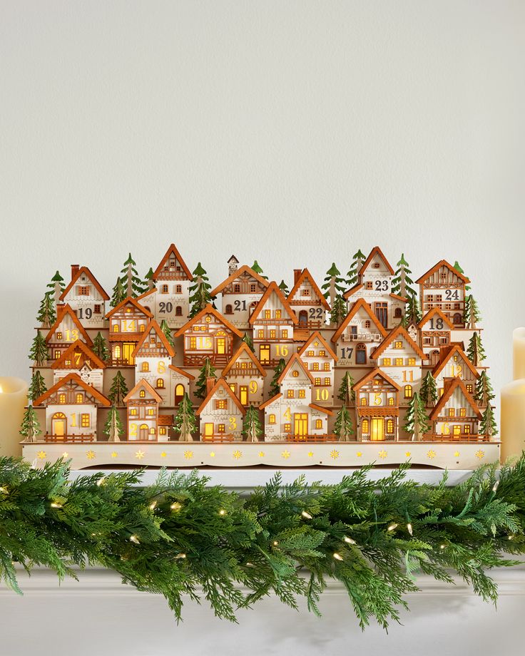 a lighted christmas village is displayed on a mantle with evergreen garland and candles in front of it