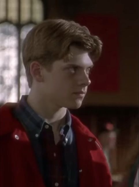 the young man is wearing a red jacket and blue shirt in front of a stained glass window