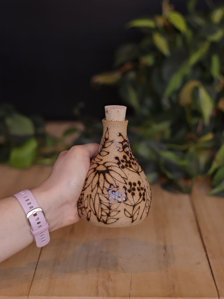 a hand holding a small vase on top of a wooden table next to a plant