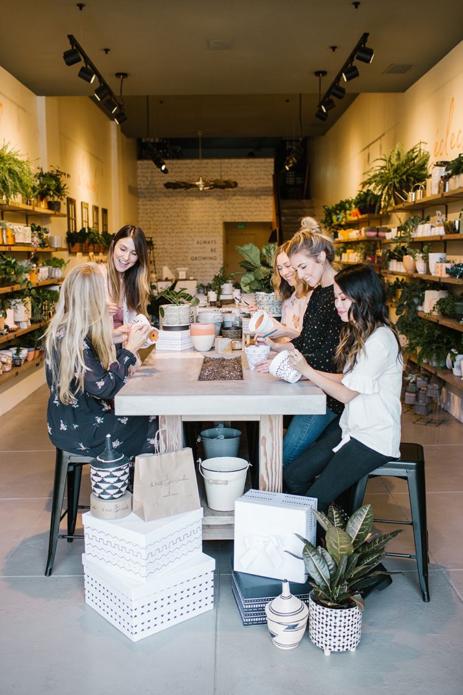 three women are sitting at a table in a flower shop and one woman is holding a cup