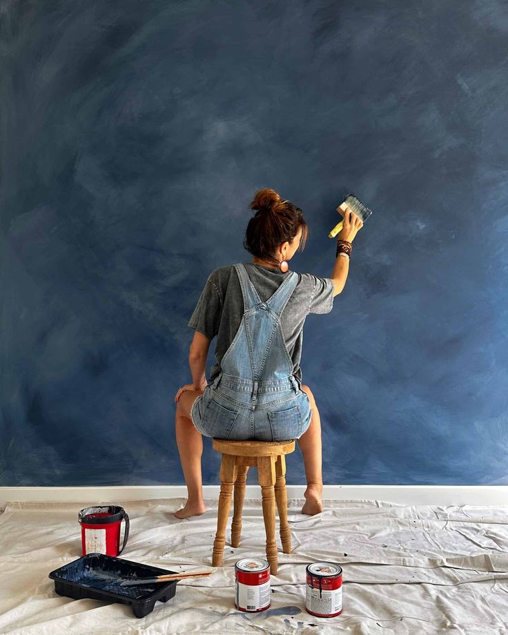 a woman sitting on top of a wooden stool painting a wall with blue chalk paint