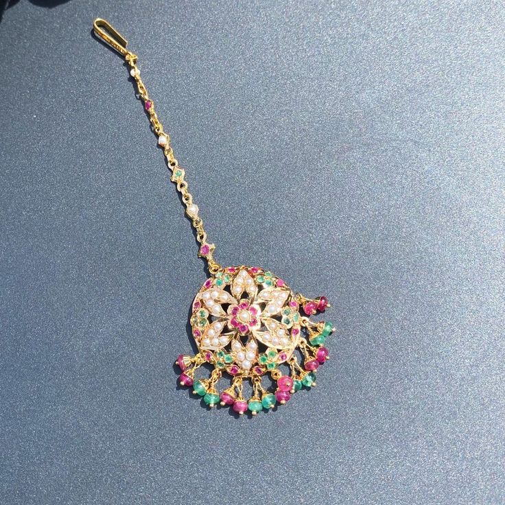 This magnificent mang teeka / tikka is designed in 22k gold with an intricately placed ruby, emerald and pearl design. Its chain measures 4 inches in length with a detachable hook at the end. It boasts an impressive total weight of 8.39 gms, 0.85 gms of which is concentrated in the ruby-emerald beads. This timeless piece is the perfect accessory for special occasions and can be passed down for generations as a treasured heirloom. The blend of precious gems and gold make it a stunning piece sure Festive Gold Plated Tikka As A Gift, Festive Temple Jewelry Tikka With Jeweled Details, Traditional Jeweled Tikka For Festive Occasions, Traditional Jeweled Tikka For Festive Season, Festive Yellow Gold Kundan Tikka, Bollywood Jeweled Tikka For Festive Occasions, Festive Bollywood Jeweled Tikka, Festive Yellow Gold Tikka With Tilla, Jeweled Temple Jewelry Tikka For Festivals