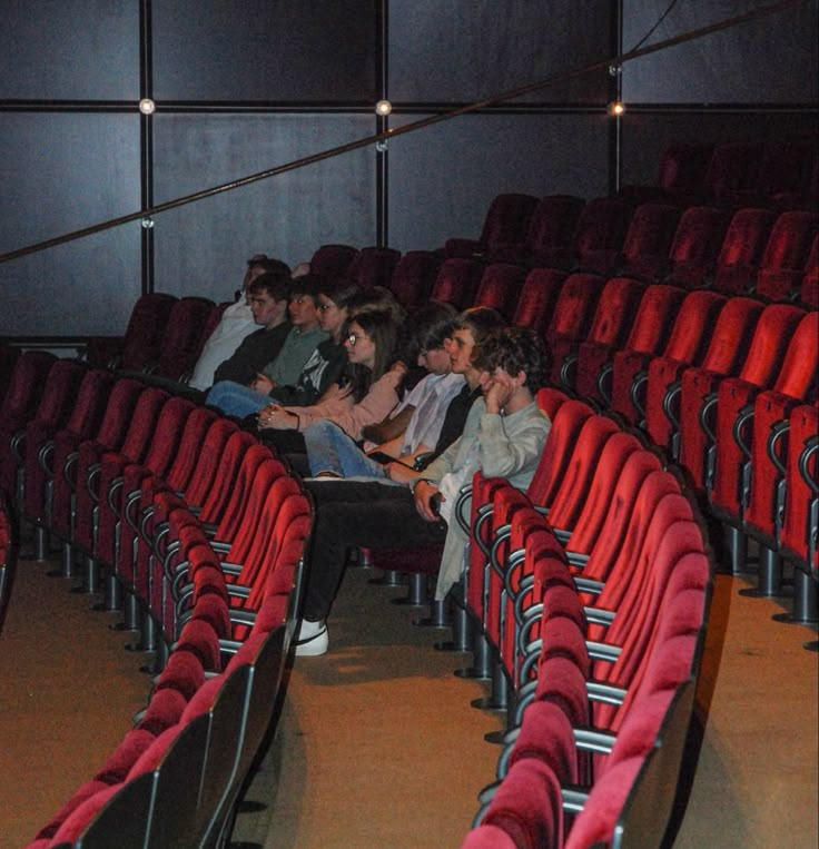 some people are sitting in an empty auditorium with red seats and one person is on his cell phone