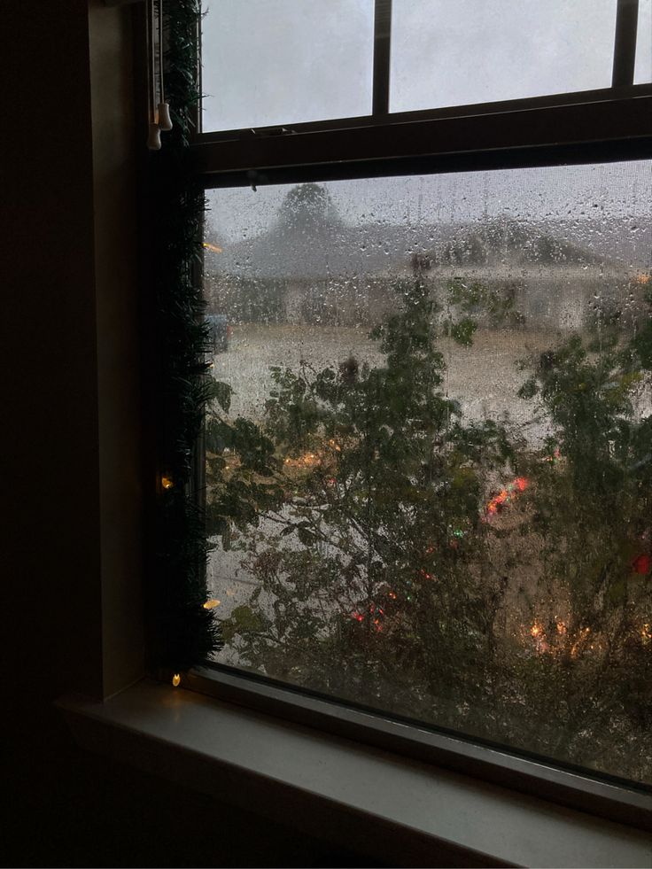 a window with rain falling on it and trees outside the window, looking out at an open field