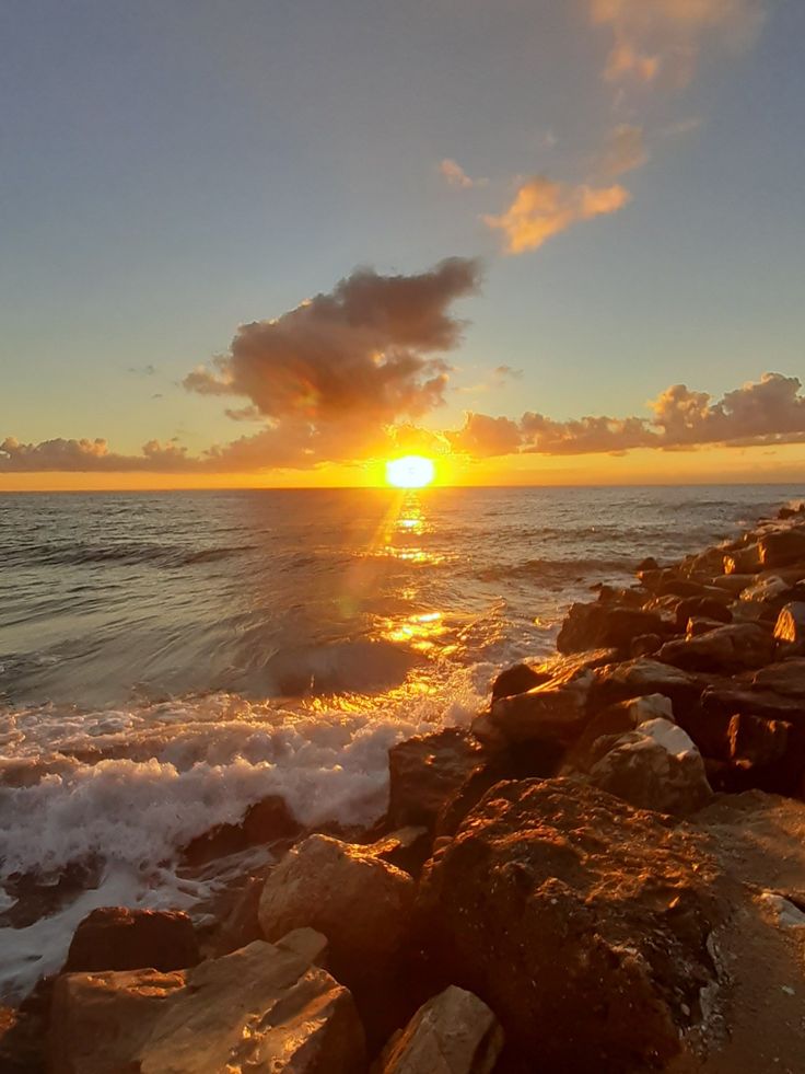 the sun is setting over the ocean with rocks on the shore and waves coming in