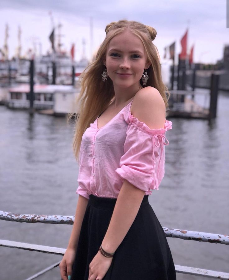 a woman standing on a dock next to the water wearing a pink shirt and black skirt