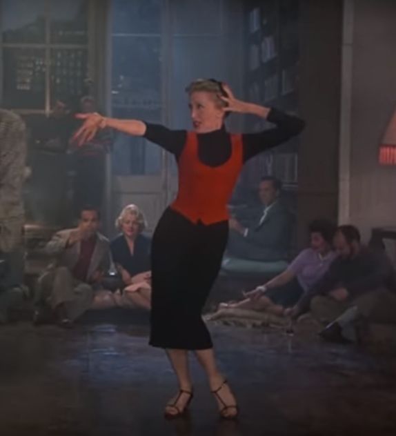 a woman in a red top and black skirt is dancing with other people sitting on the floor