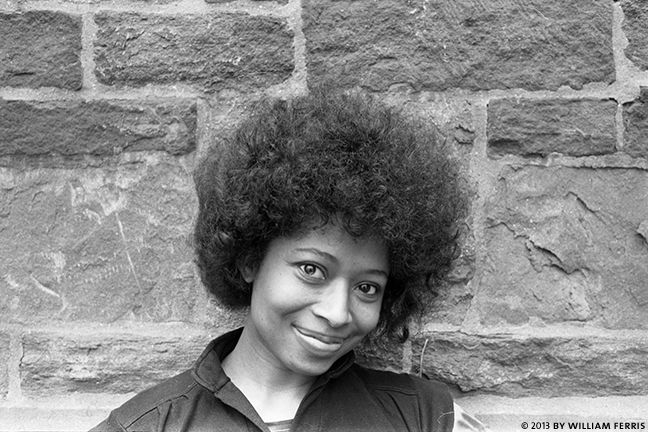 a black and white photo of a woman with an afro smiling at the camera, in front of a brick wall