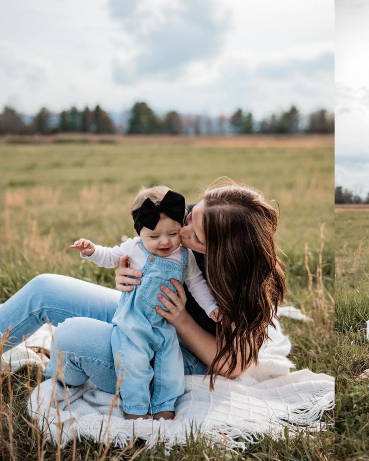 a woman holding a baby while sitting on top of a blanket in the middle of a field