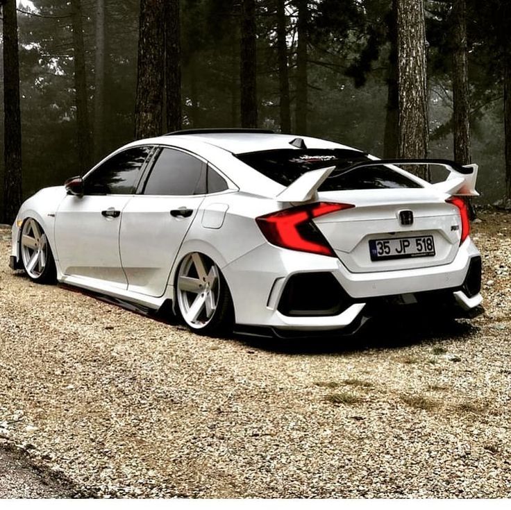 the rear end of a white car parked on top of a dirt road in front of trees