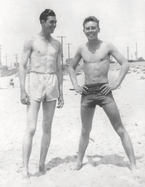 two men standing next to each other on top of a sandy beach in swim trunks