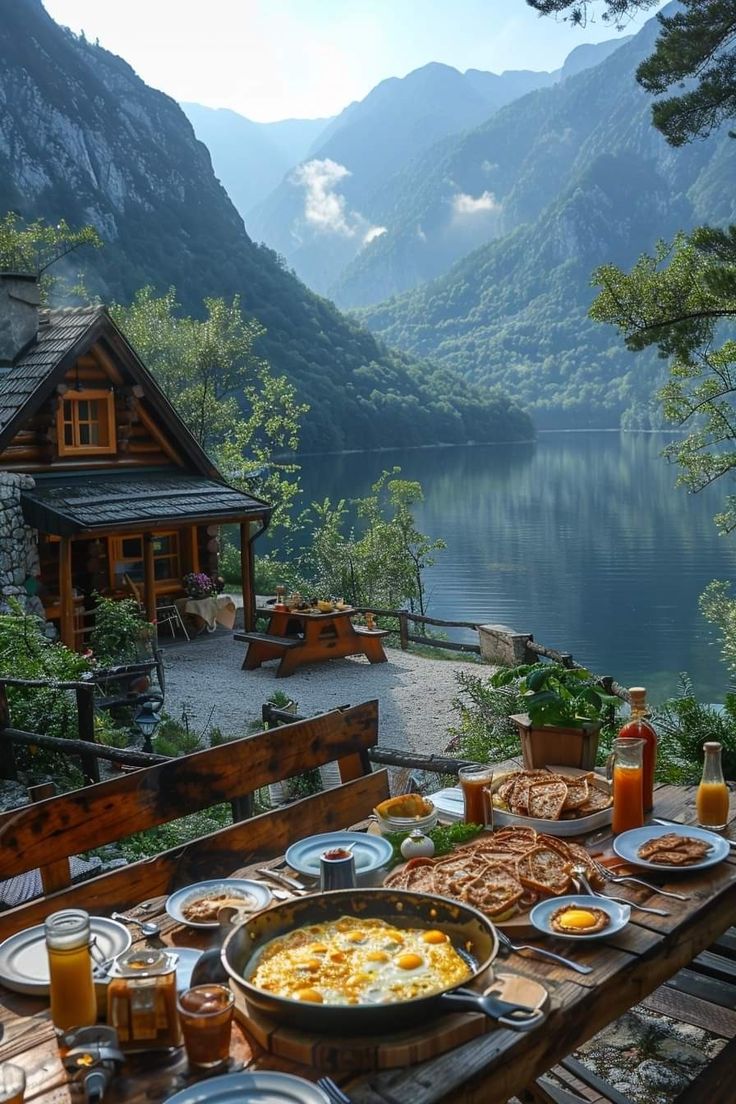 a wooden table topped with plates of food next to a body of water and mountains