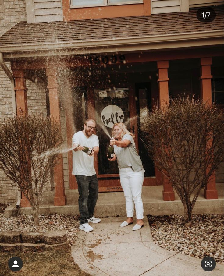 a man and woman standing in front of a house spraying water on each other with their hands