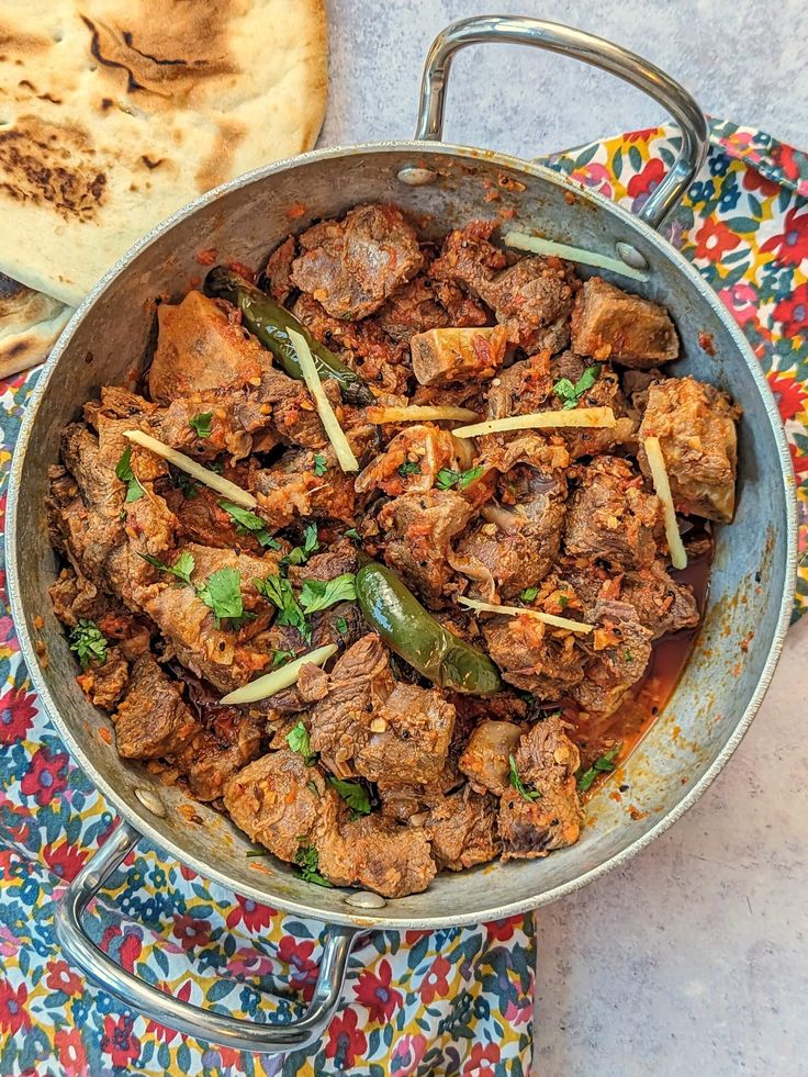 a pan filled with meat and vegetables on top of a colorful cloth next to bread