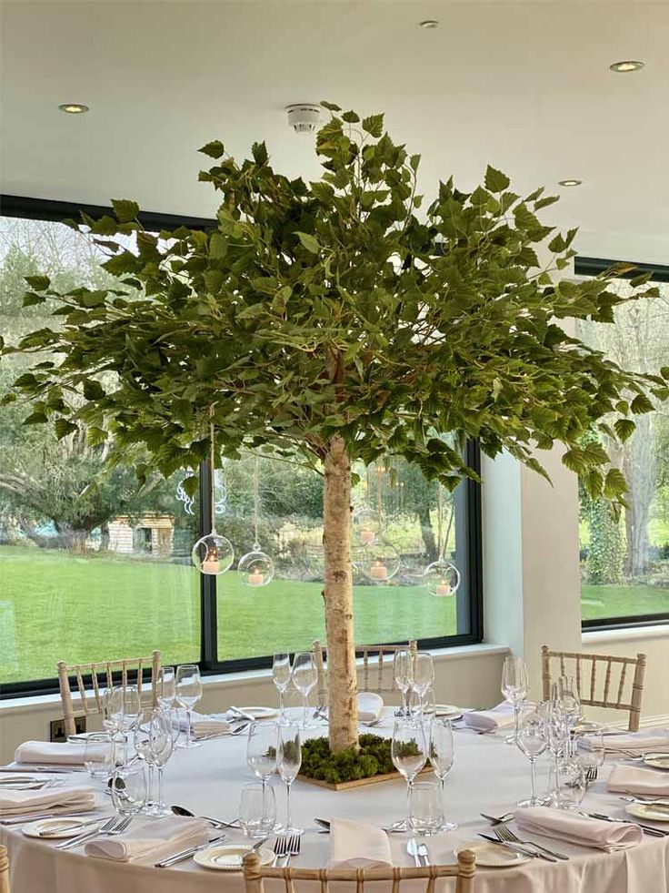 a table set up for a formal function with a tree in the center and chairs around it