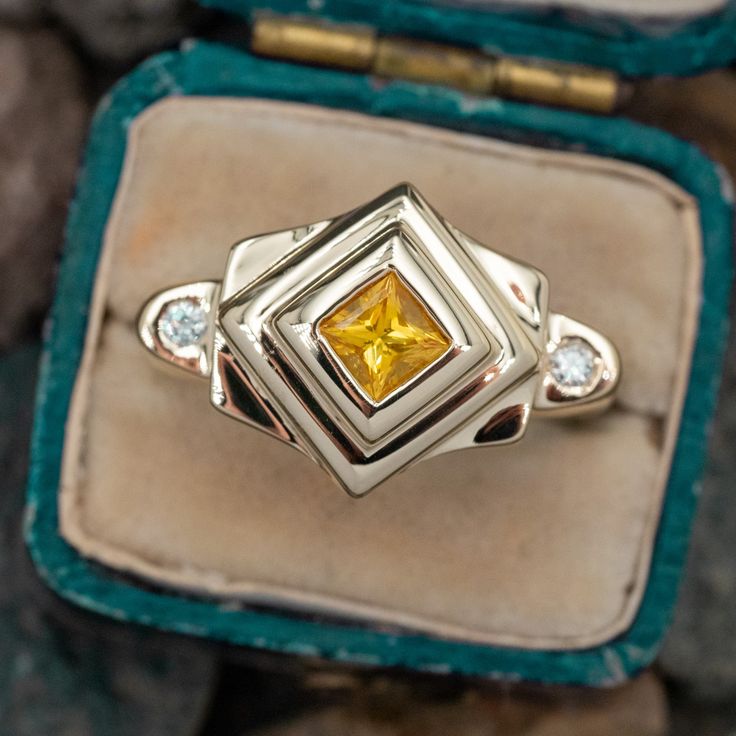 This intriguing estate ring holds a yellow sapphire center stone with a round brilliant cut diamond accent set on either side. The ring has a weighty design with a Euro shank and is currently a size 7. Yellow Sapphire Ring Brilliant Cut Fine Jewelry, Yellow Brilliant Cut Sapphire Ring, Yellow Sapphire Ring With Center Stone In Yellow Gold, Anniversary Yellow Sapphire Ring Brilliant Cut, Yellow Diamond Ring In 14k Gold, 14k Gold Yellow Diamond Ring, Yellow Sapphire Ring With Diamond, Yellow 14k Gold Diamond Ring, Yellow Sapphire Ring With Round Cut Gemstone