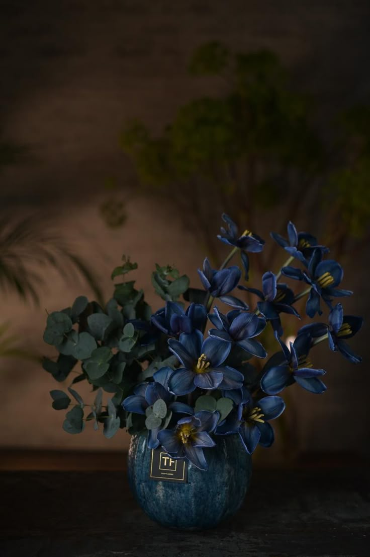 a blue vase filled with purple flowers on top of a wooden table next to a potted plant