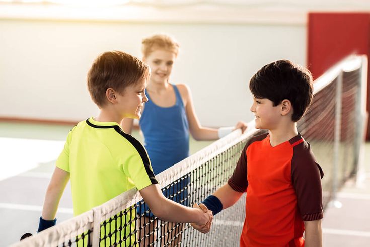 two young boys shaking hands over the net of a tennis court while another boy looks on