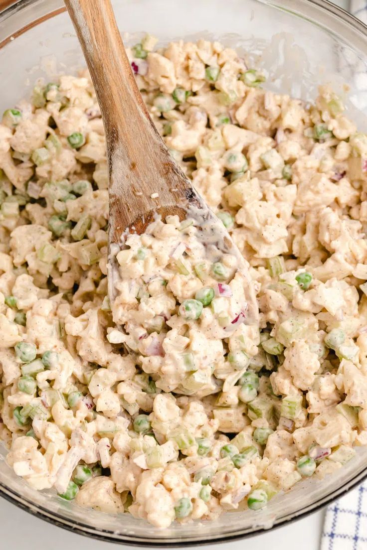 a wooden spoon in a glass bowl filled with chicken salad and peas on a blue checkered table cloth