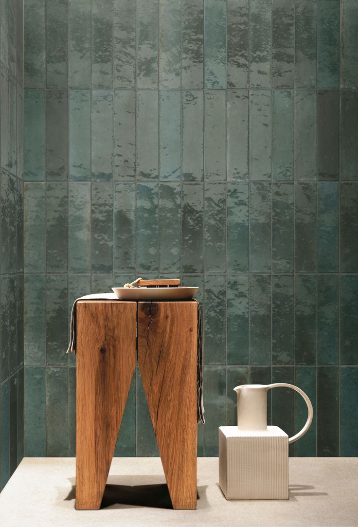 a wooden table sitting next to a white vase on top of a counter in front of a green tiled wall