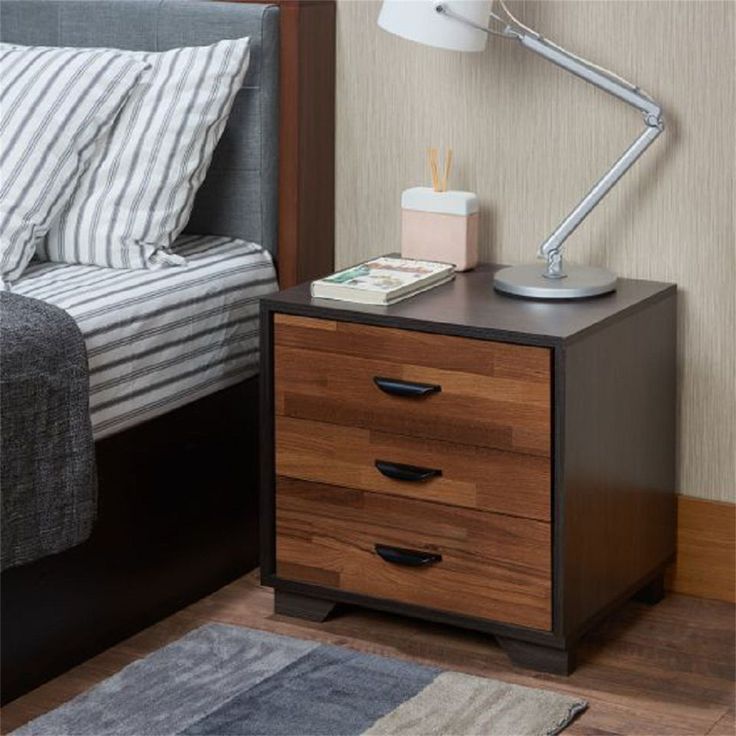 a nightstand with two drawers and a lamp next to it on top of a bed