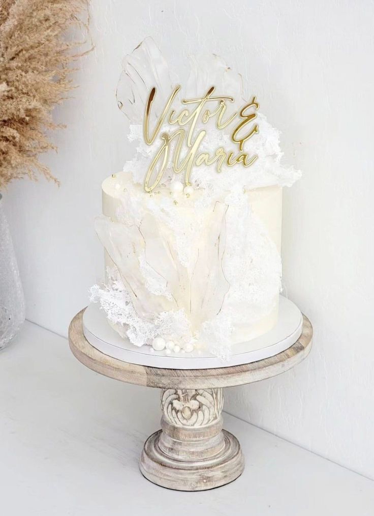 a white cake with gold lettering sitting on top of a table next to dried flowers