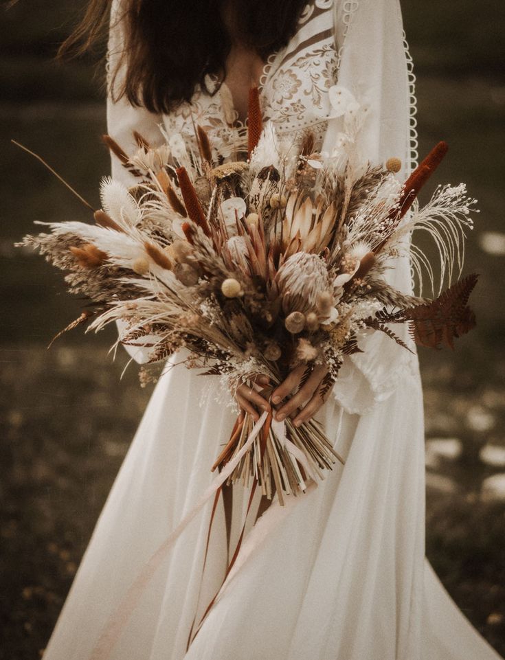 a woman in a white wedding dress holding a bouquet of dried flowers and feathers on her arm