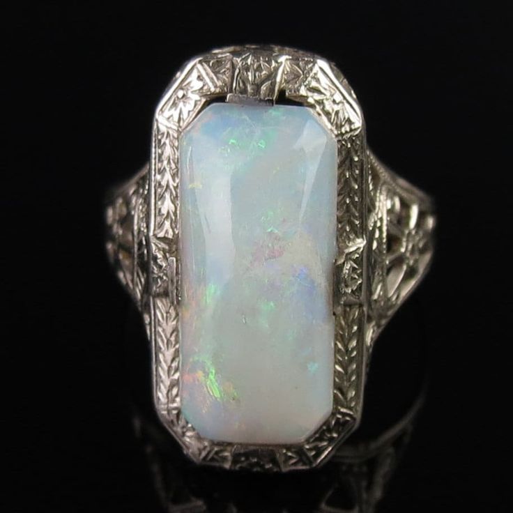 A natural white opal tops this ornate 14k white gold ring by famed jewelers, Ostby & Barton. A carved Art Deco design frames the 13mm x 6mm x 3.8mm cabochon. The white opal has a pinkish undertone with subtle flashes of greens and reds. A pierced filigree design with scrollwork and floral elements decorates the tapered shoulders. The inside of the ring is signed, "OB" (hallmark attributed to Ostby & Barton) followed by "14k" (white gold). The ring measures a petite size 4 (a pinky size for some) Silver Cabochon Platinum Rings, Art Deco White Gold Jewelry With Cabochon, Silver Platinum Rings With Cabochon, Platinum Silver Rings With Cabochon, Platinum Rings With Silver Cabochon, Vintage Opal Jewelry For Anniversary, Classic Opal Moonstone Anniversary Ring, Classic Moonstone Opal Ring For Anniversary, Heirloom White Opal Ring