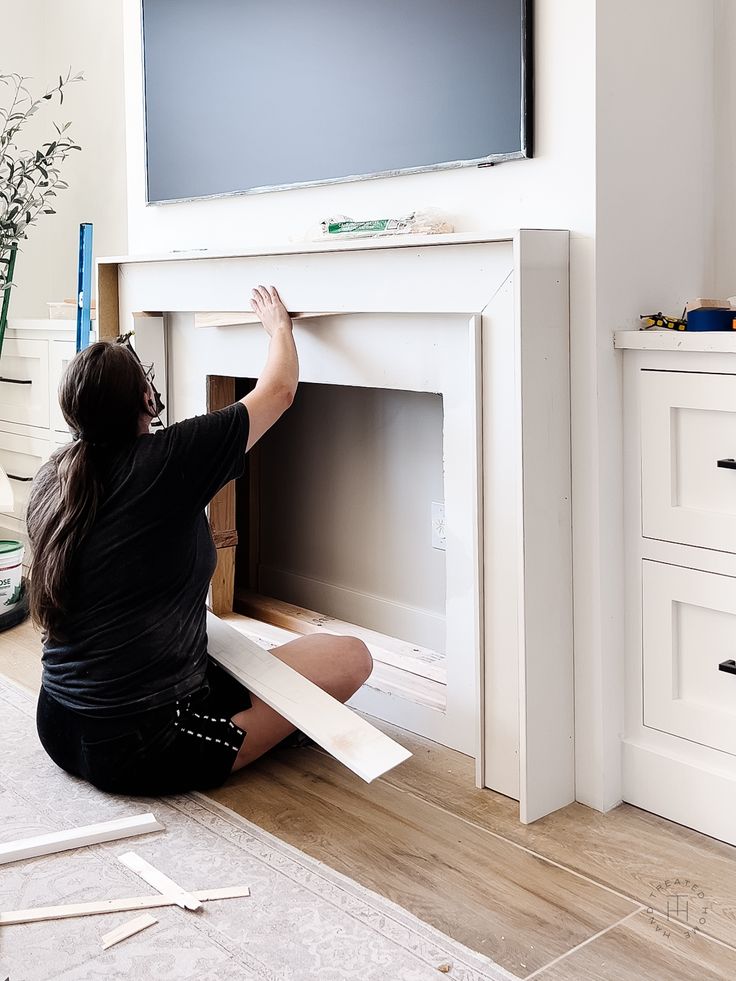 a woman sitting on the floor in front of a fireplace with a flat screen tv above it