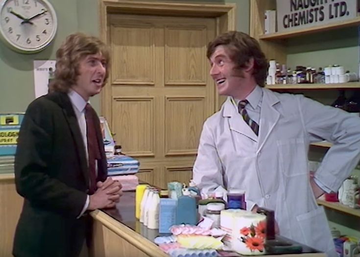 two men in white lab coats talking to each other at a counter with various items on it