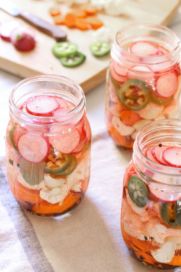 four jars filled with pickles and vegetables on top of a white cloth next to a cutting board