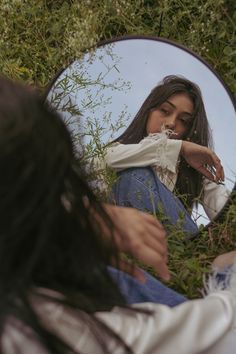 a woman sitting in the grass looking at her reflection in a round mirror with long hair