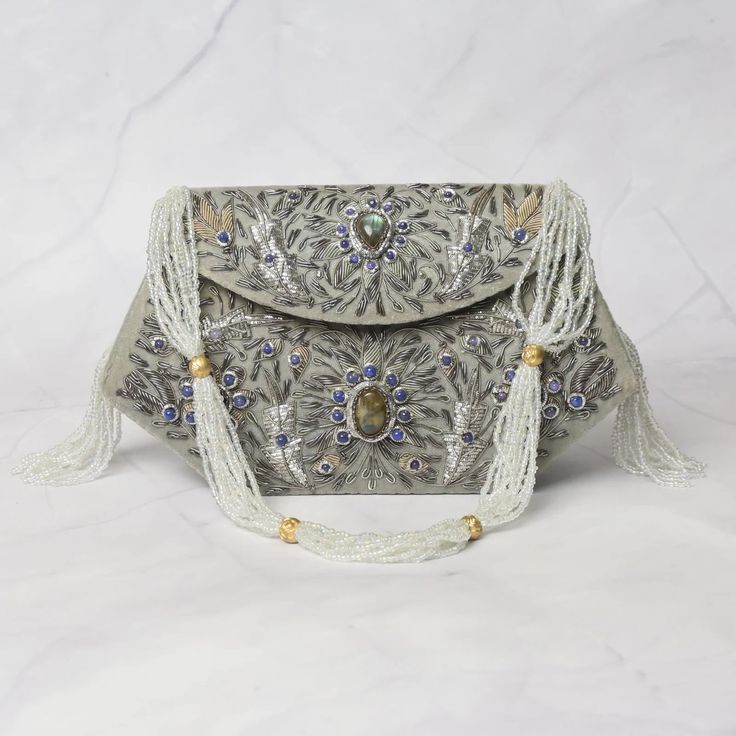 Step into the world of sophistication and charm with our "Grey Velvet Elegance" Boat Shape Clutch. This exquisite accessory is the epitome of opulence and artistry, designed for those who appreciate fine craftsmanship. Size(cm): 22*14*7 Material: Velvet Fabric & Metal Thread Stones: Labradorite & Onyx Base Color: Grey Pockets: Single Handmade with meticulous care, this clutch features sumptuous grey velvet adorned with intricate Zardozi work. The floral motifs come to life with the richness of Labradorite and Onyx gemstones. Labradorite, known for its iridescent play of colors, adds a mystical allure to the clutch. Onyx, with its deep grey hue, exudes timeless elegance. The boat shape design provides ample space to carry your essentials in style. The velvety texture feels luxurious to the Luxury Gray Shoulder Bag For Formal Occasions, Luxury Beaded Shoulder Bag, Luxury Handmade Shoulder Bag, Luxury Gray Evening Bag, Gray Luxury Shoulder Bag For Evening, Elegant Engraved Gold Bags, Hand Embellished Silver Bag For Formal Occasions, Formal Hand Embellished Silver Bag, Formal Silver Hand-embellished Bag