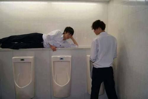 two young men standing next to urinals in a bathroom, one leaning on the wall