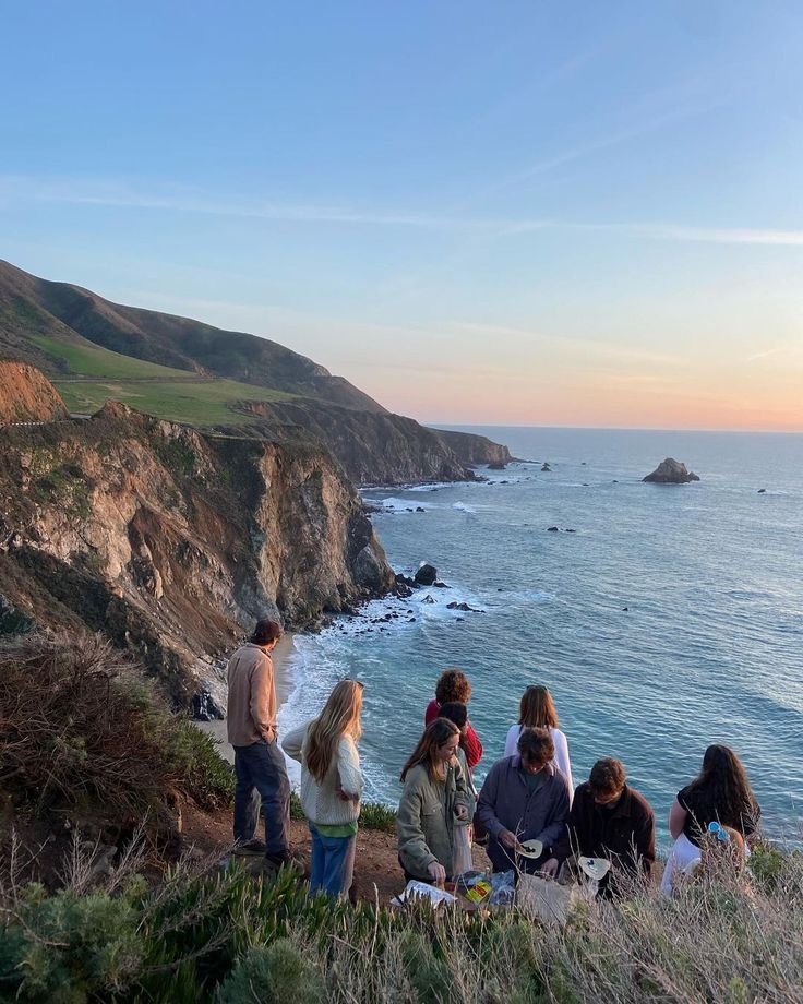 group of people sitting on the edge of a cliff overlooking the ocean and cliffs at sunset