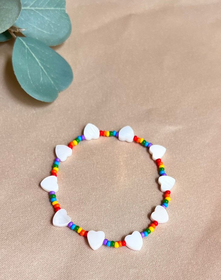 a bracelet with white hearts and rainbow colored beads on a beige surface next to a green leaf