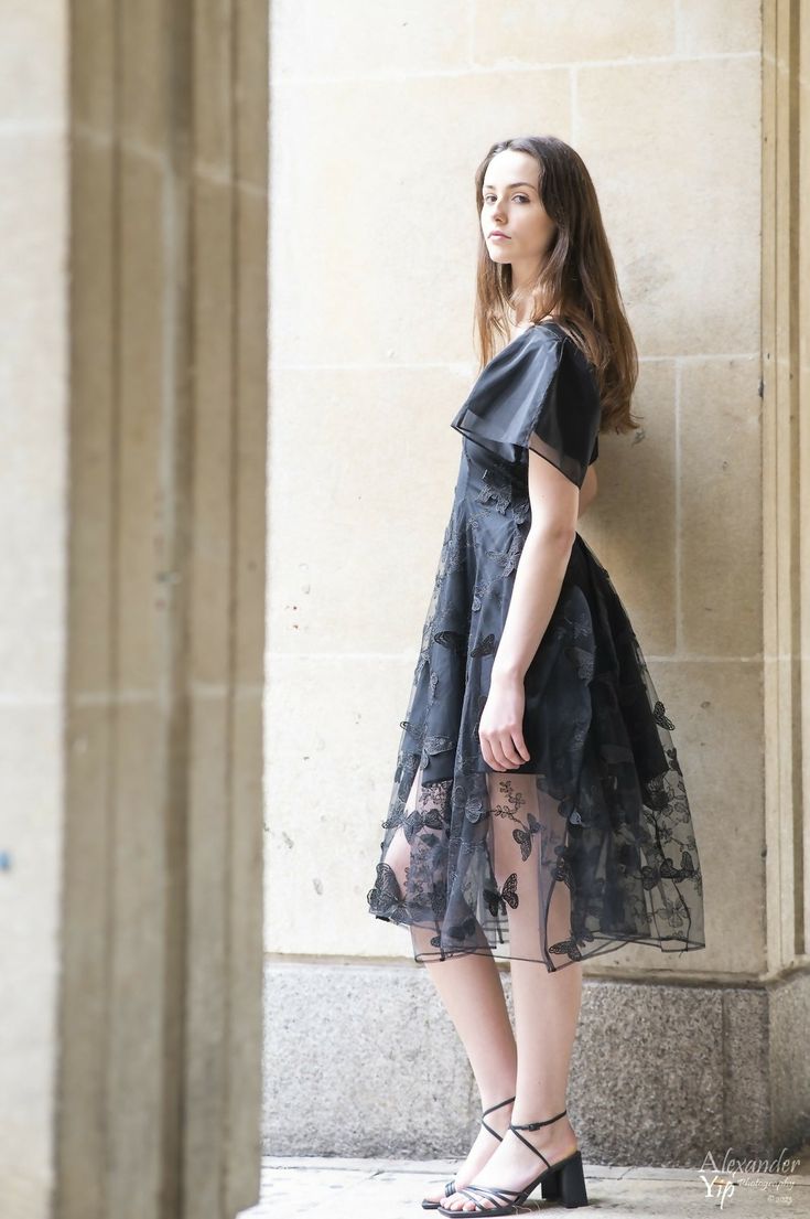 a woman standing in front of a stone wall wearing black dress and high heeled sandals