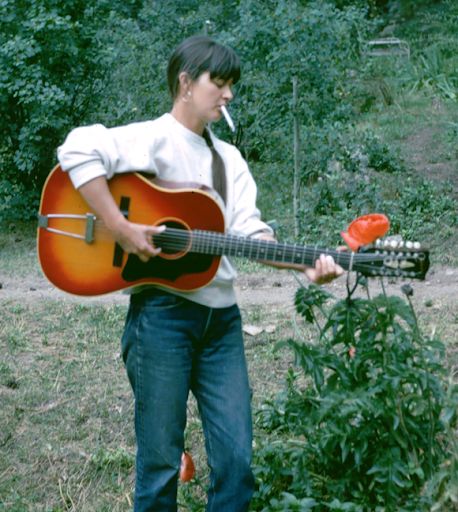 a man holding an acoustic guitar in front of a bush with trees and bushes behind him