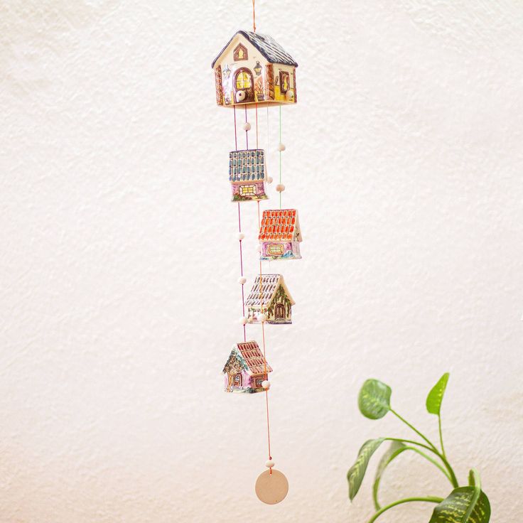 a house shaped mobile hanging from a wall next to a potted plant and white wall