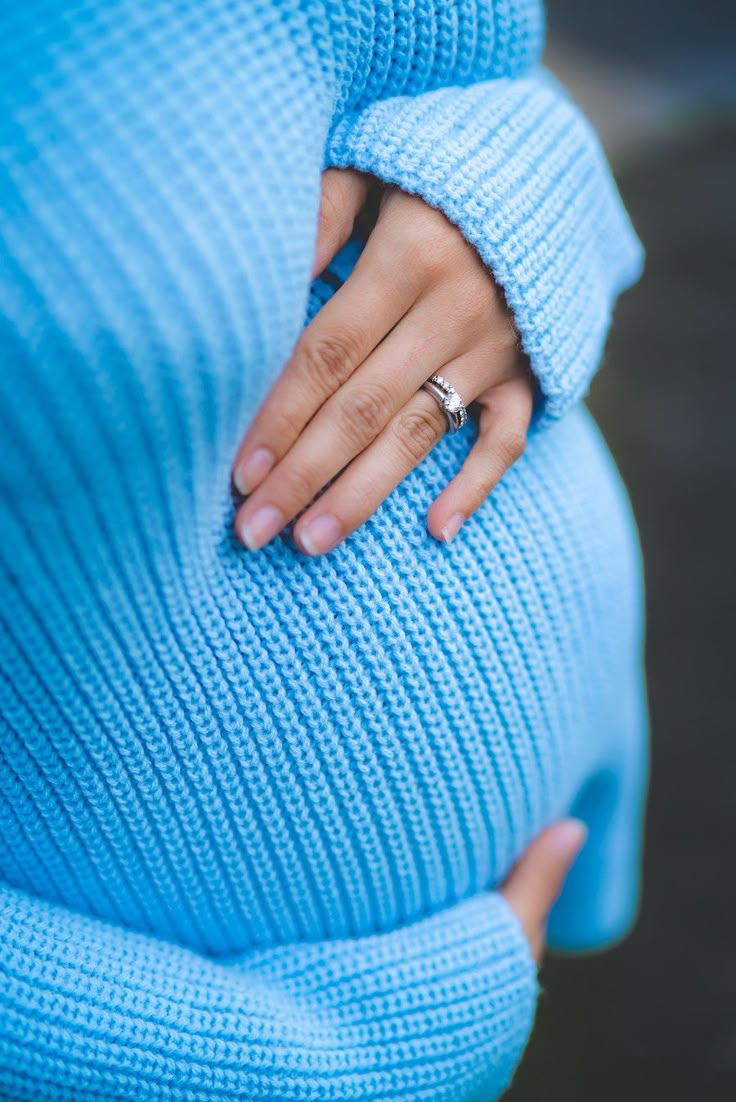 a woman's hand on her stomach wearing a blue sweater and matching engagement ring