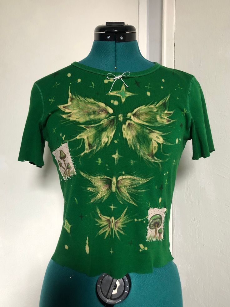 Bleach-painted kelly green T-shirt with butterflies, mushrooms, and stars graphic. Patches are adhered with fabric glue and sewn on. Small bow detail on the collar. All graphic details are done in fabric marker. Should fit sizes XS-S. This piece is handmade, handpainted, one of a kind, and made from a reused T-shirt. Wash inside out or carefully handwash in cold water.  In case of accidents, free repair is offered. Price is calculated based on hours spent making the product (minimum wage), effor Green Graphic Print Y2k Tops, Y2k Green Graphic Print Tops, Green Y2k Graphic Print Tops, Fitted Fairy Grunge T-shirt For Spring, Spring Fairy Grunge Fitted T-shirt, Fairy Grunge Fitted T-shirt For Spring, Fitted Green Graphic Print T-shirt, Fitted Green T-shirt With Graphic Print, Fairy Grunge Fitted Short Sleeve T-shirt