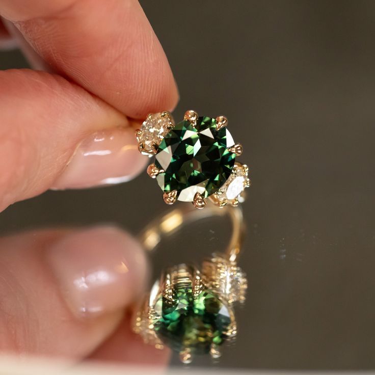 a close up of a person's hand holding a ring with a green and white diamond