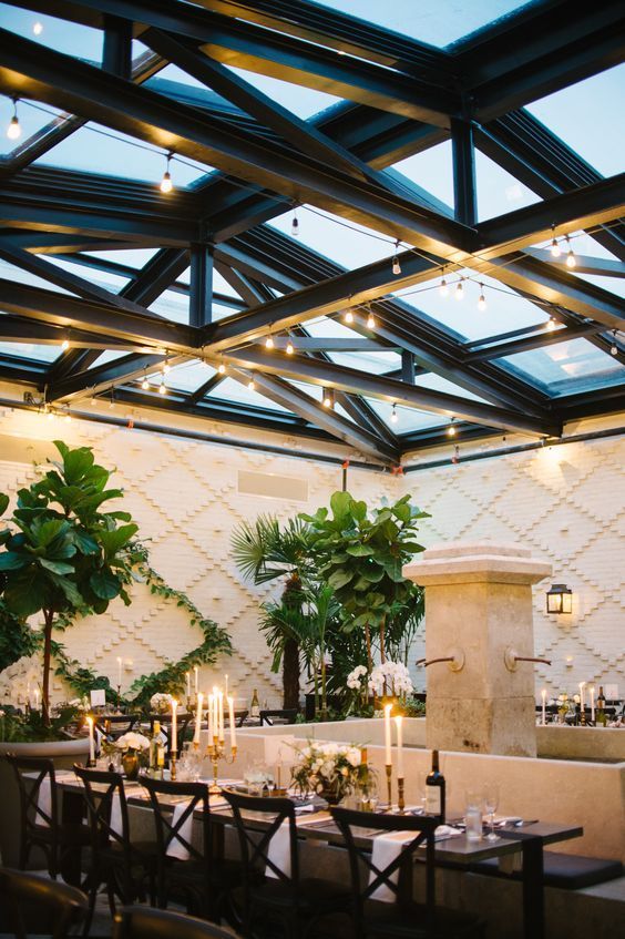 an indoor dining area with tables, chairs and potted plants in the center is lit by candles