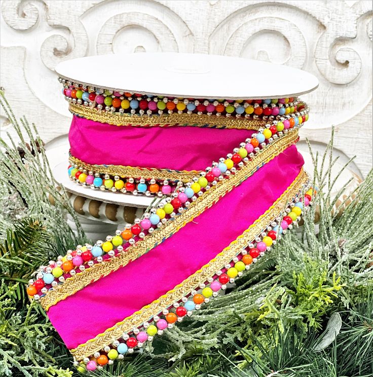 a pink and gold ribbon with beaded trim on top of evergreen branches in front of a white wall